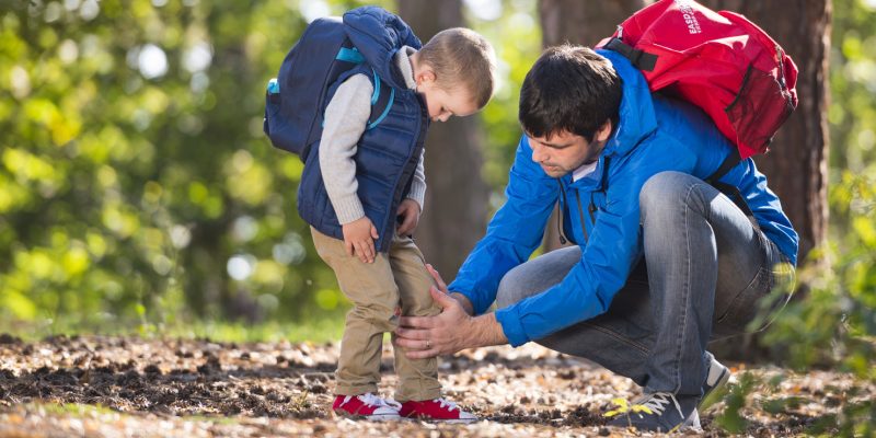 Photograph showing a young boy with his dad in the woods. Dad is crouching down checking the boys left leg.