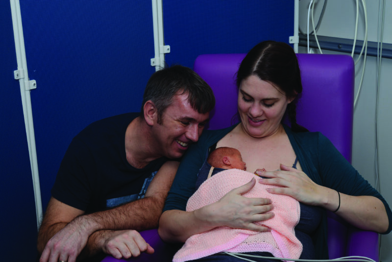 image of mother and father, mother sat on chair with baby in skin to skin, both parents smiling